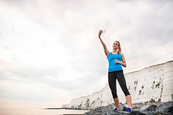 Young sporty woman runner with earphones and smartphone on the beach, taking selfie. Copy space.