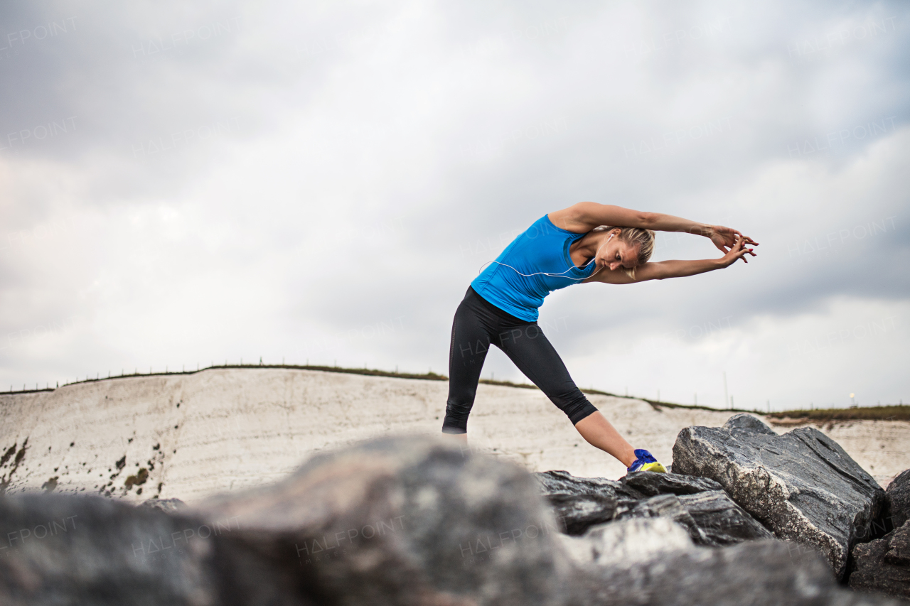 Young sporty woman runner with earphones standing outside on the rocks by beach in nature, listening to music and stretching. Copy space.