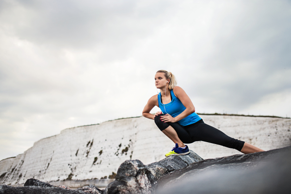 Young sporty woman runner with earphones standing outside on the rocks by beach in nature, listening to music and stretching. Copy space.