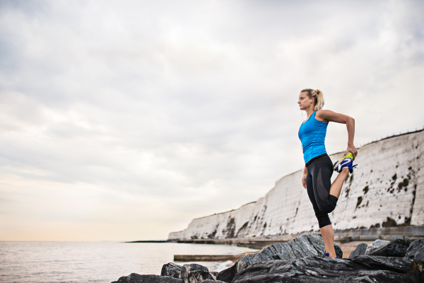 Young sporty woman runner with earphones stretching outside on the beach in nature. Copy space.