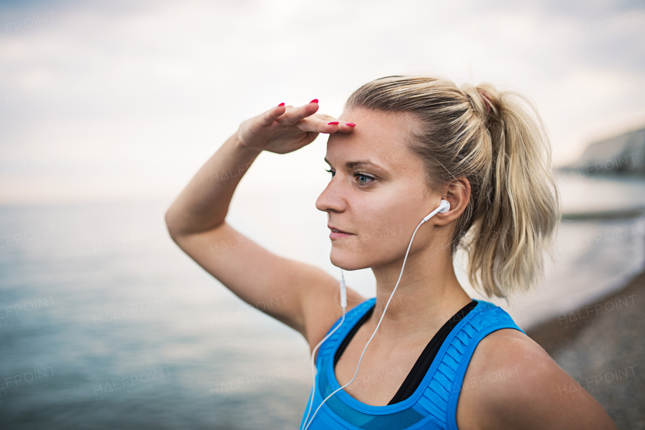 Young sporty woman runner with earphones standing on the beach outside, looking far away with her hand at the forehead.