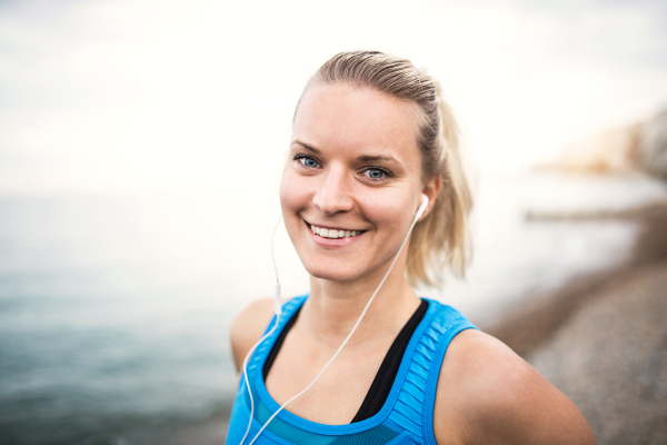 Young sporty woman runner with earphones standing outside on the beach in nature, listening to music and resting. Copy space.