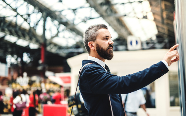 Side view of hipster businessman standing on bus or train station, checking timetable.