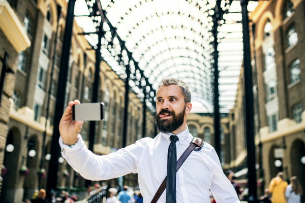 Hipster businessman with smartphone standing on the trian station in London, grimacing and taking selfie.