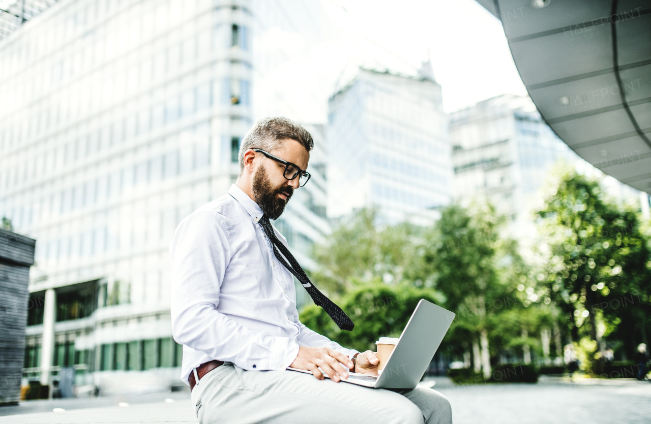 Hipster businessman with laptop sitting outdoors on the street in the city.