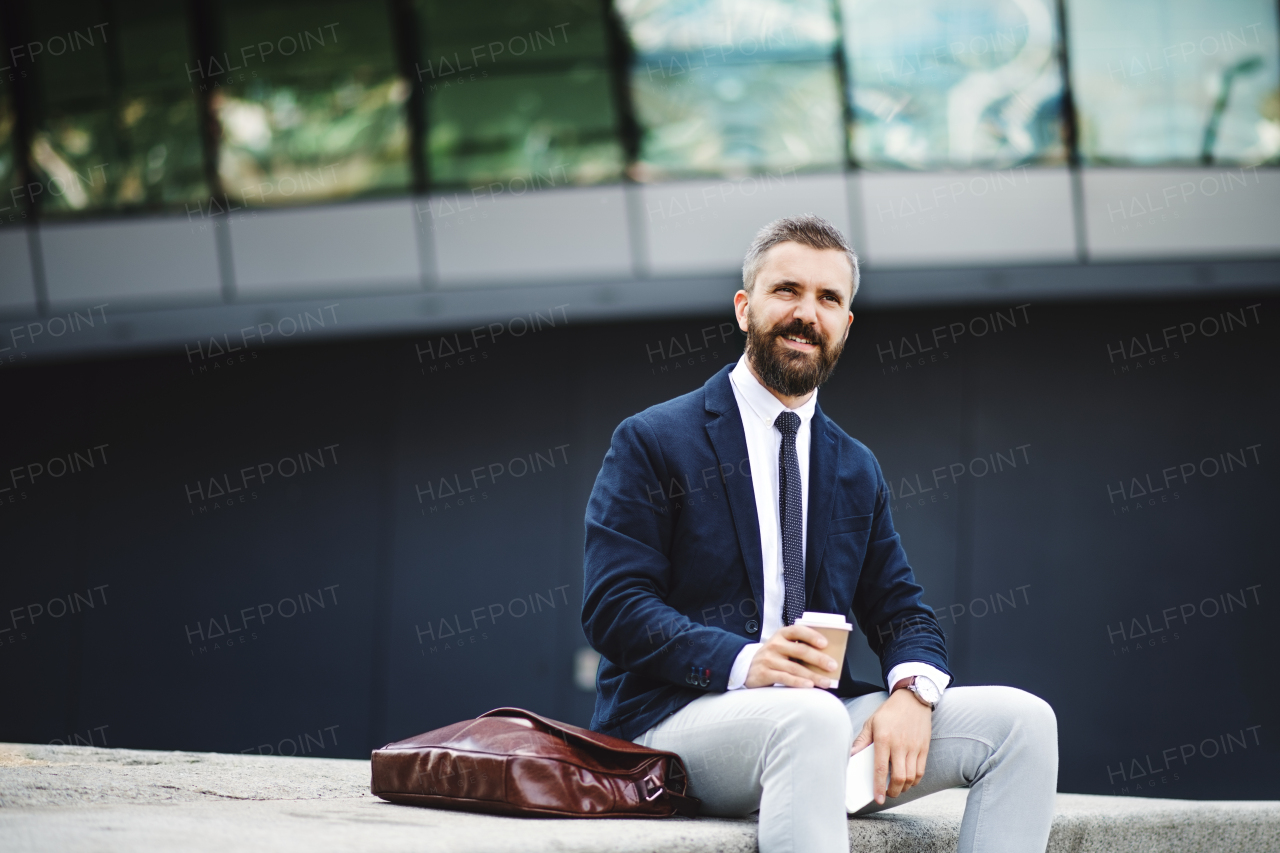 Serious hipster businessman with smartphone, laptop bag and coffee cup sitting outdoors in the city.