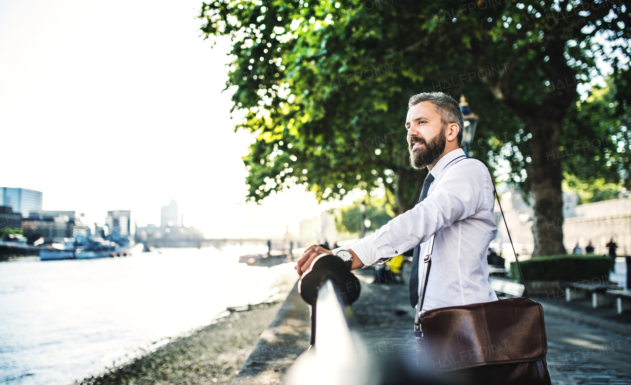 Happy hipster businessman with laptop bag standing by the river in London, holding onto a railing.