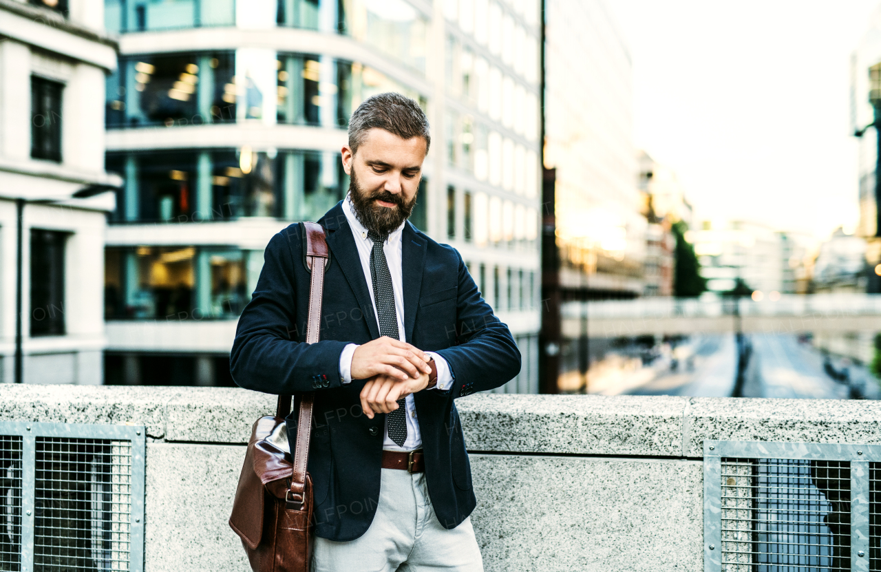 Hipster businessman with laptop suitcase standing on the bridge in city, checking the time.