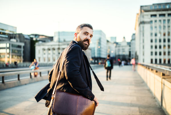 A rear view of hipster businessman with a bag running on the street in the city, looking back.