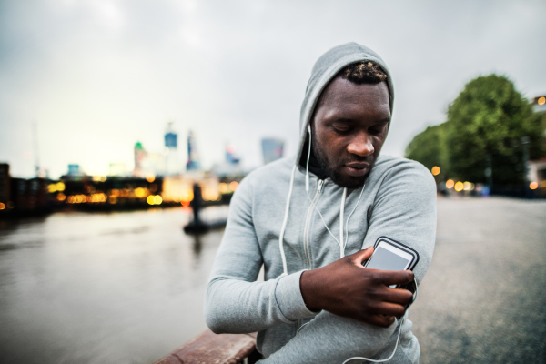 Young sporty black man runner with smartwatch, earphones and smartphone in an armband on the bridge in a city, resting.