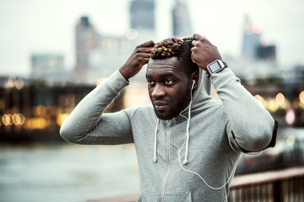 Young sporty black runner with smartwatch, earphones and smartphone in an armband on the bridge in a city, resting. A man putting a hood on head.