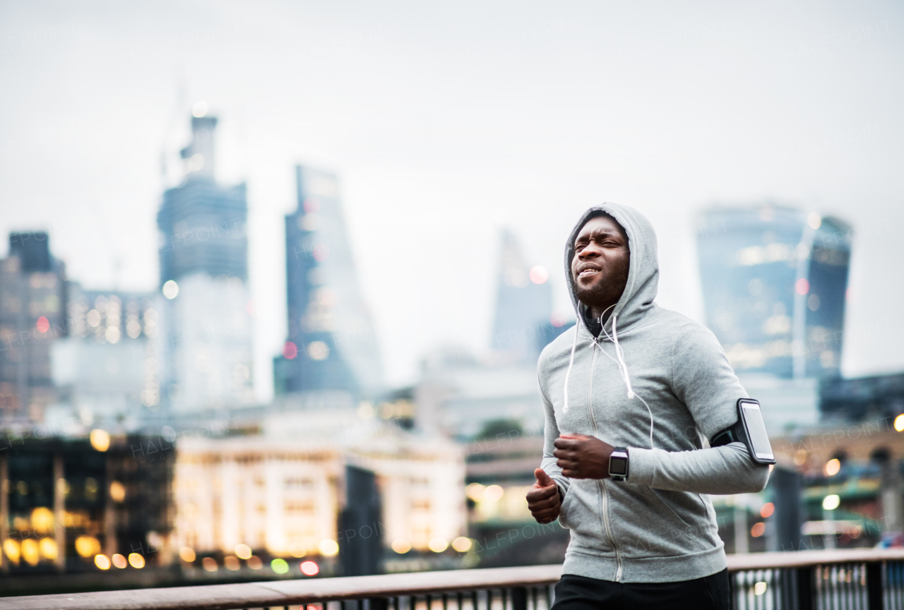 Young sporty black man runner with smartwatch and earphones running on the bridge outside in a city.