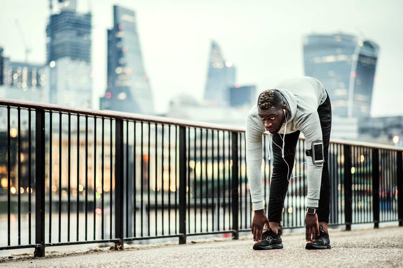 Young sporty black man runner with smartwatch, earphones and smartphone in an armband on the bridge in a city, stretching.