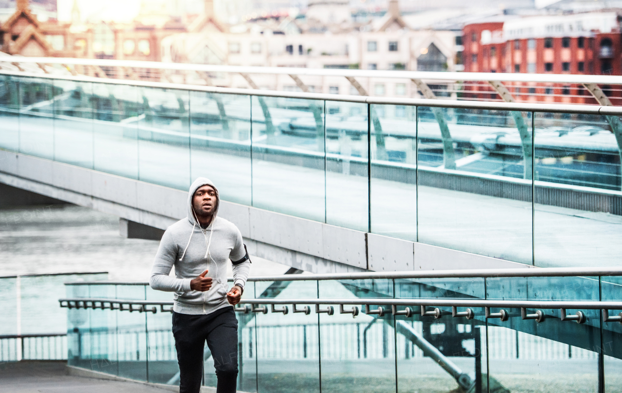 Young sporty black man runner with smartwatch and earphones running on the bridge outside in a city.