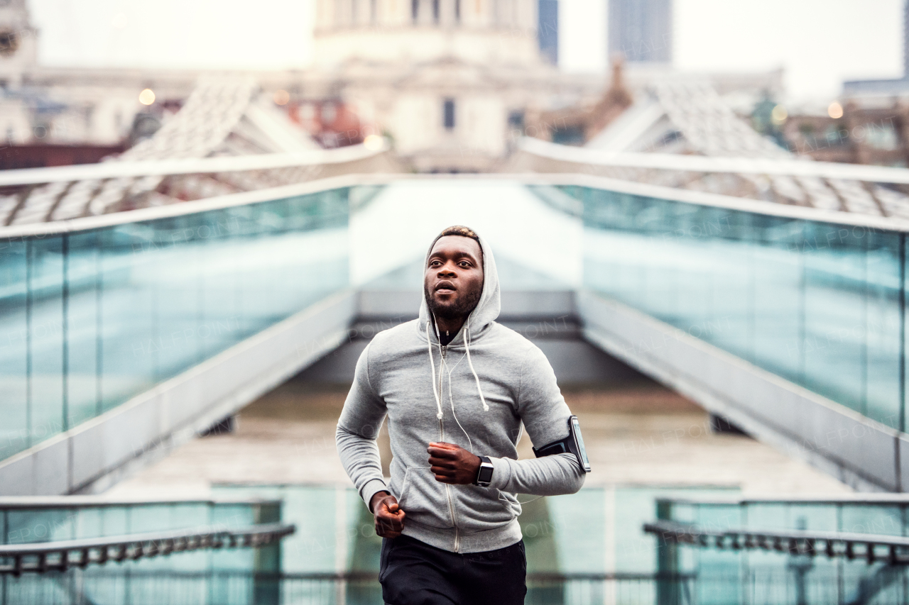 Young sporty black man runner with smartwatch and earphones running on the bridge outside in a city.