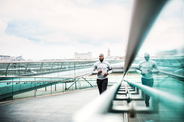 Young sporty black man runner with smartphone in armband and earphones running on the bridge outside in a city. Copy space.