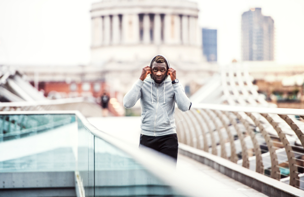 Young sporty black man runner with smartwatch, earphones and smartphone in an armband on the bridge in a city, resting.