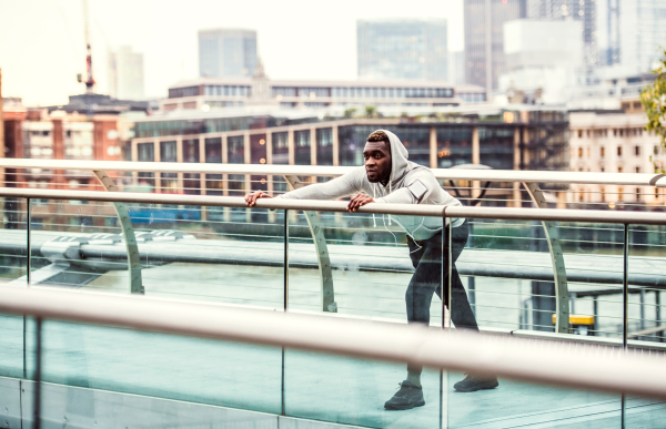 Young sporty black man runner with smartwatch, earphones and smartphone in an armband on the bridge in a city, stretching.