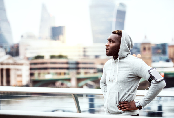 Young sporty black man runner with smartwatch, earphones and smartphone in an armband on the bridge in a city, resting. Copy space.