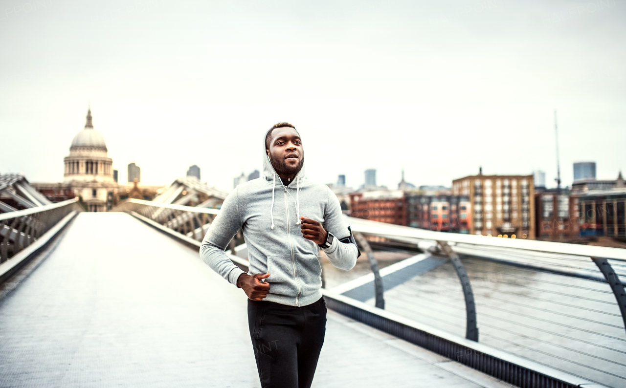 Young sporty black man runner with smartwatch and earphones running on the bridge outside in a city.