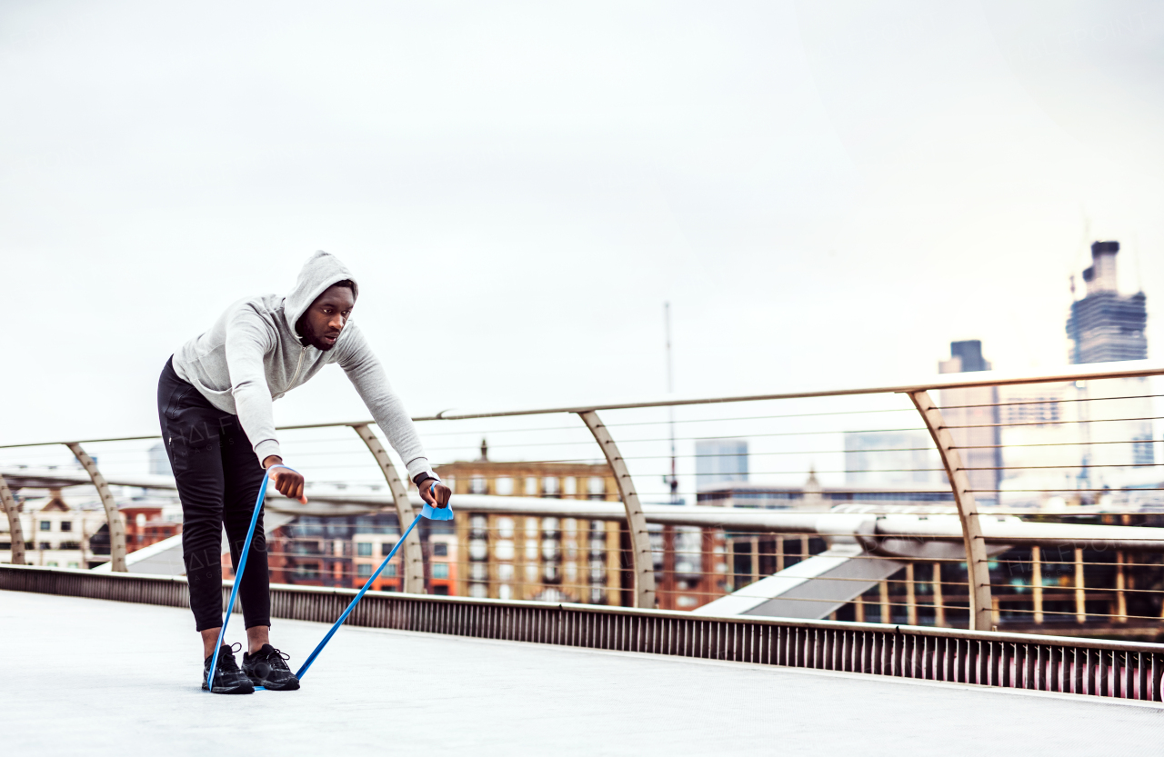 Young active black sportsman exercising with elastic rubber bands on the bridge in London.