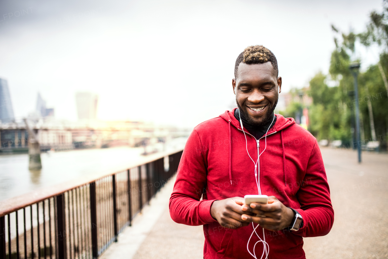 Young sporty black man runner with earphones, smartwatch and smartphone on the bridge outside in a city, listening to music.