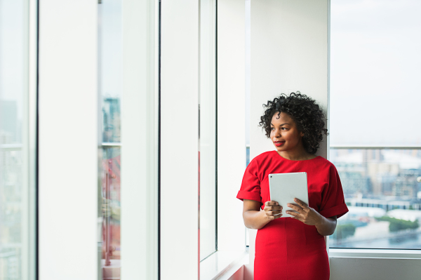 A portrait of woman with tablet standing by the window against London panorama. Copy space.