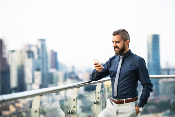 A portrait of businessman with smartphone standing against London rooftop view panorama, text messaging. Copy space.