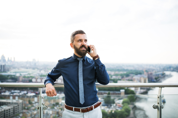 A portrait of businessman with smartphone standing against London rooftop view panorama, making a phone call. Copy space.