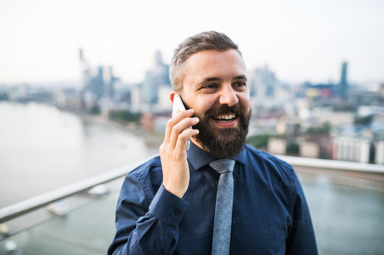 A portrait of businessman with smartphone standing against London rooftop view panorama, making a phone call.