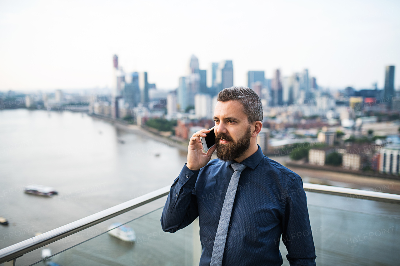 A portrait of businessman with smartphone standing against London rooftop view panorama, making a phone call. Copy space.