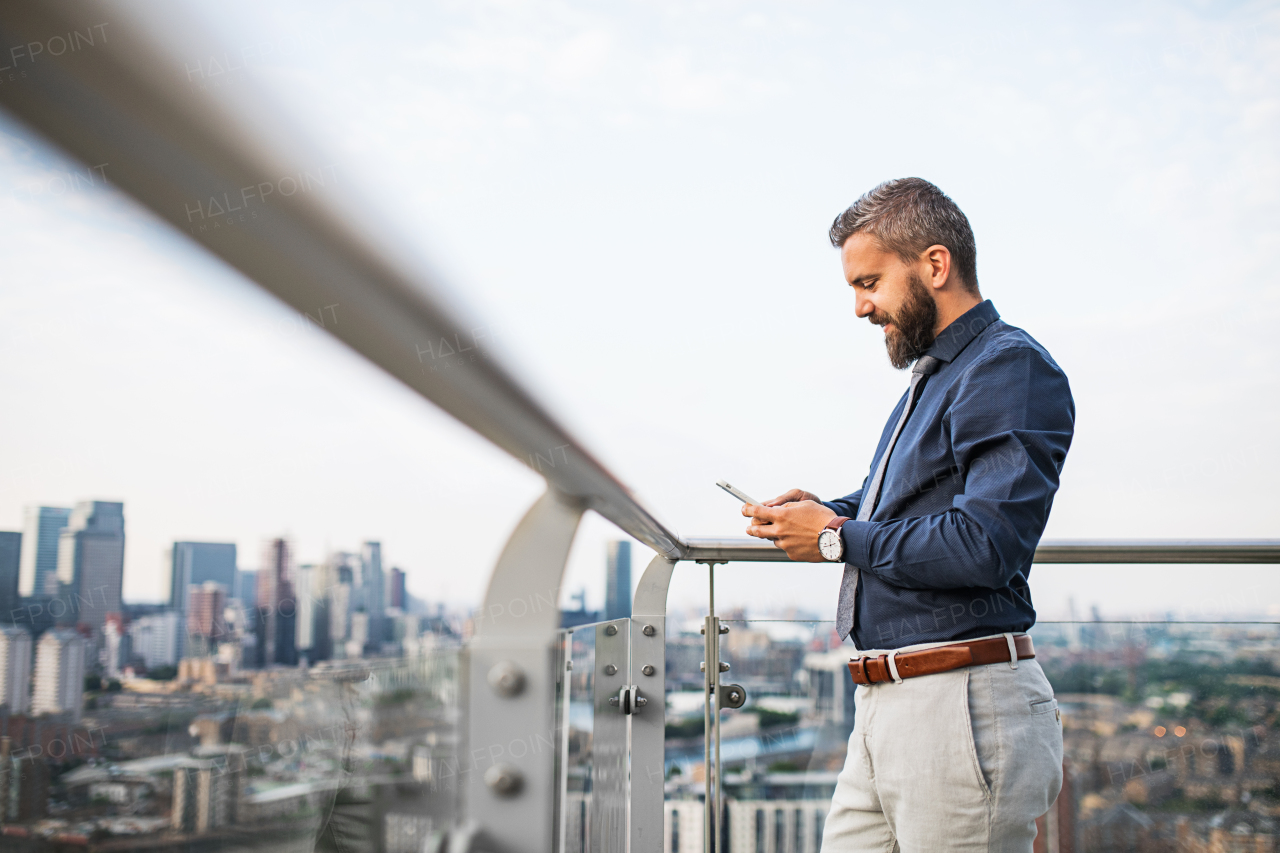 A portrait of businessman with smartphone standing against London rooftop view panorama, texting. Copy space.