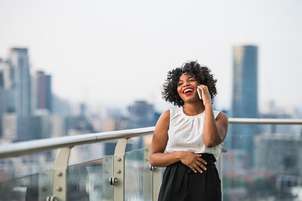 A portrait of happy black businesswoman standing against London rooftop view panorama, making a phone call. Copy space.