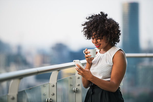 A portrait of a black woman with smartphone standing on a terrace, drinking coffee. Copy space.