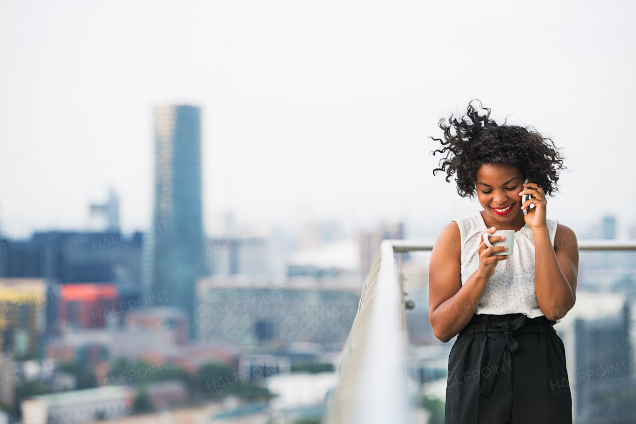 A portrait of a woman with coffee and smartphone standing on a terrace, making a phone call. Copy space.