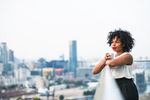 A portrait of a black woman standing on a terrace, holding a cup of coffee. Copy space.