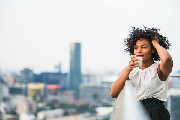 A close-up portrait of a black woman standing on a terrace, drinking coffee. Copy space.