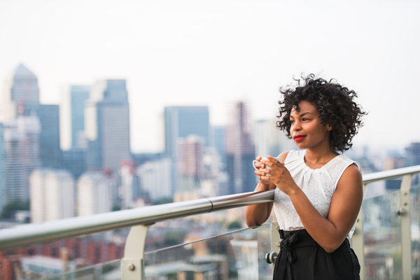 A portrait of a black woman standing on a terrace, holding a cup of coffee. Copy space.