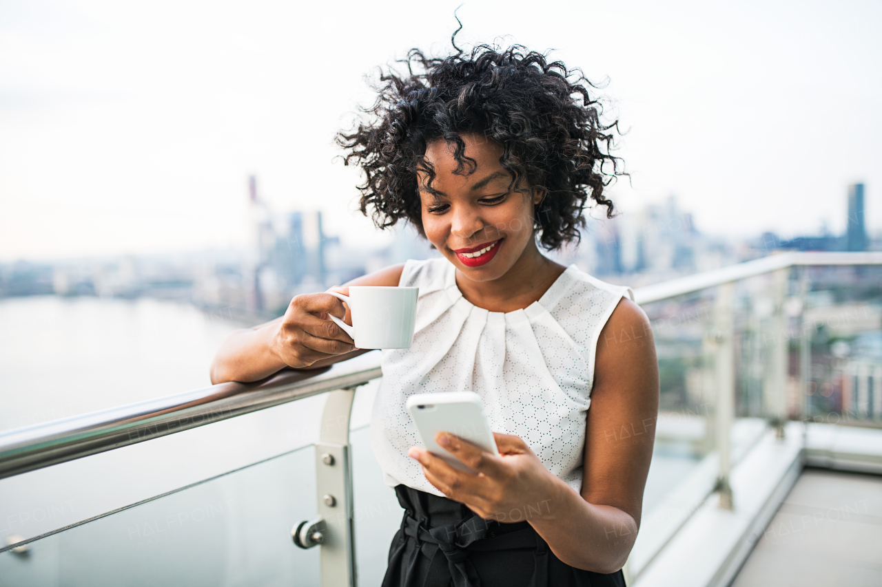 A businesswoman with coffee and smartphone standing against London view panorama, text messaging.