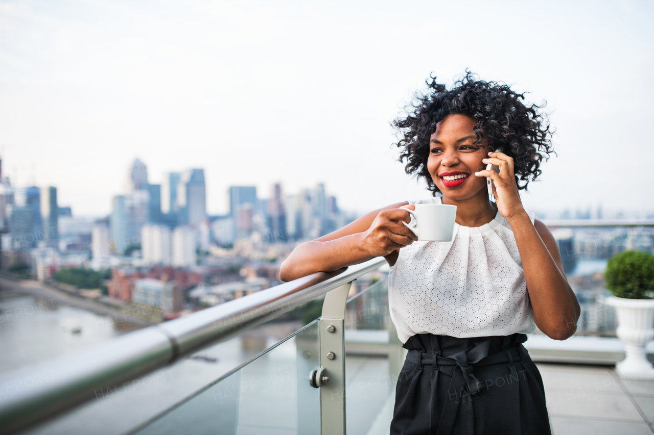 A portrait of black businesswoman with a cup of coffee standing against London rooftop view panorama, making a phone call. Copy space.