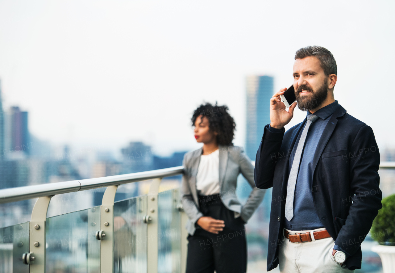 A portrait of businessman standing with a colleague in the background on a terrace against London rooftop view panorama, making a phone call.