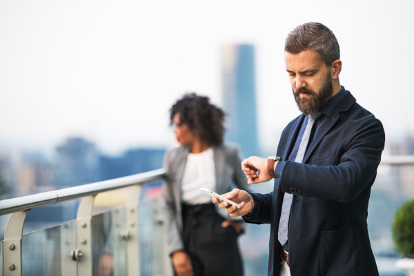 A portrait of businessman standing with a colleague in the background on a terrace against London rooftop view panorama, holding smartphone and checking the time.