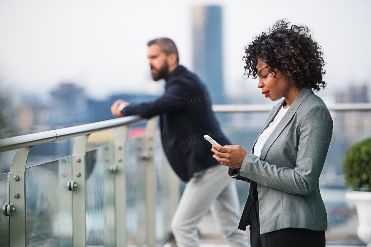 A portrait of black businesswoman standing with a colleague in the background on a terrace against London rooftop view panorama, holding smartphone.