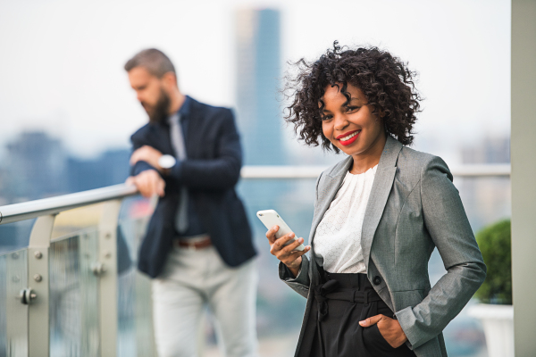 A portrait of black businesswoman standing with a colleague in the background on a terrace against London rooftop view panorama, holding smartphone.