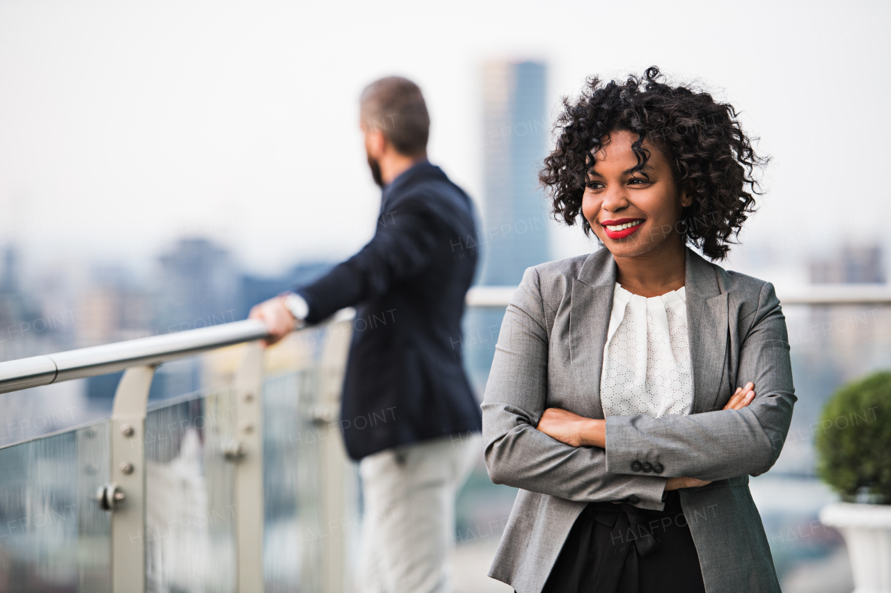 A portrait of black businesswoman standing with a colleague in the background on a terrace against London rooftop view panorama, arms corssed. Copy space.