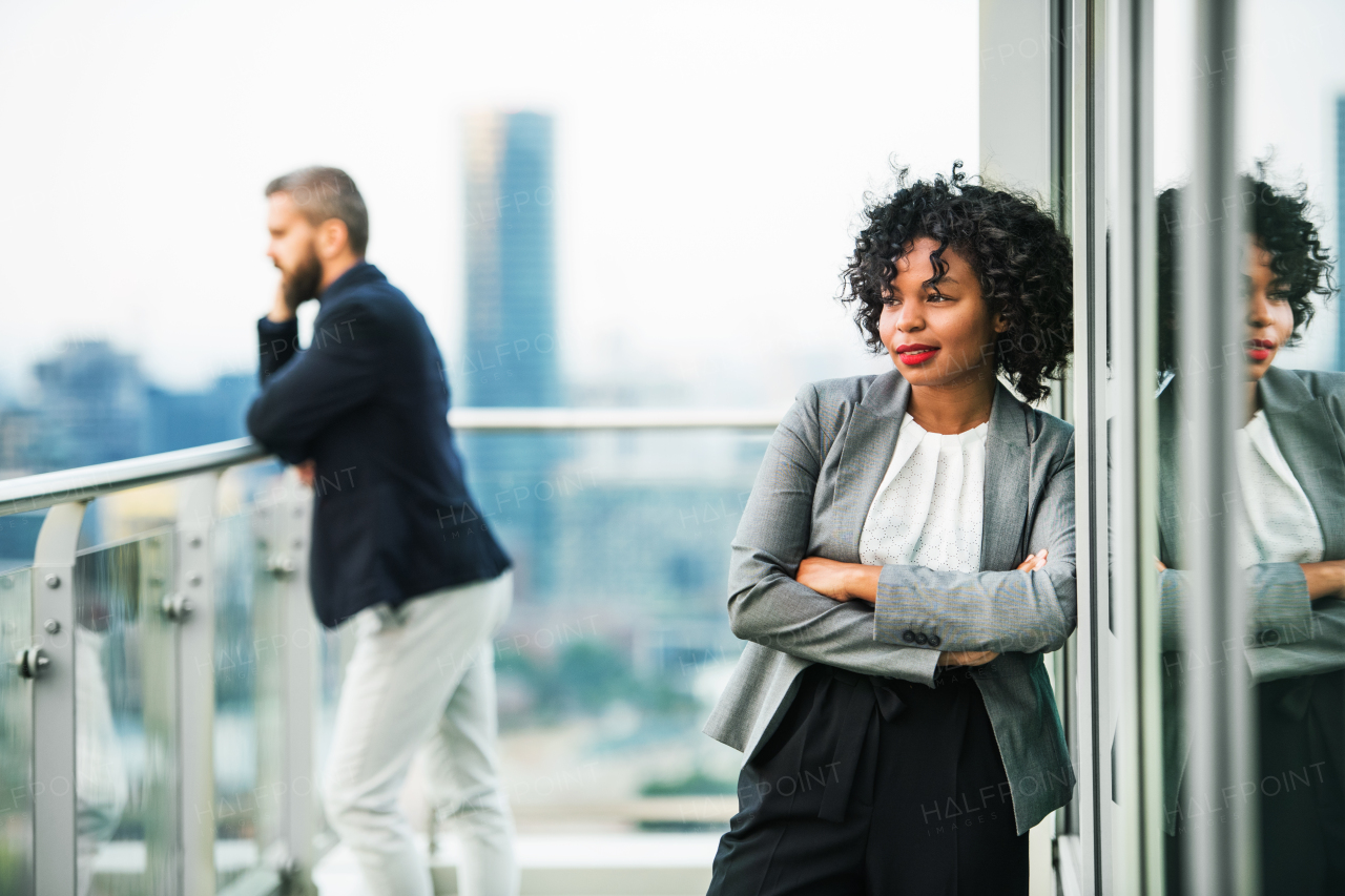 A portrait of black businesswoman standing on a terrace arms crossed, a male colleague in the background. Copy space.