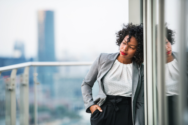 A portrait of black businesswoman standing on a terrace against London rooftop view panorama, hand in pockets. Copy space.