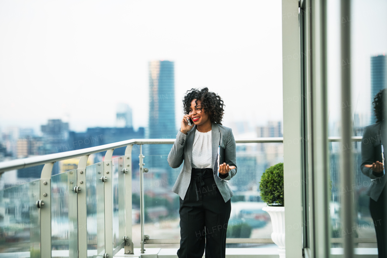 A portrait of black businesswoman with smartphone standing on a terrace against London rooftop view panorama, making a phone call. Copy space.