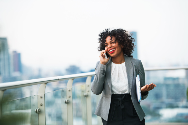 A portrait of black businesswoman with smartphone standing on a terrace against London rooftop view panorama, making a phone call. Copy space.