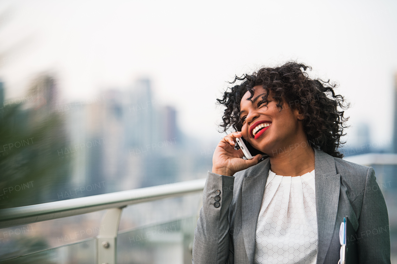 A portrait of black businesswoman with smartphone standing on a terrace against London rooftop view panorama, making a phone call. Copy space.